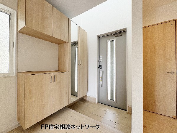 【玄関】  白を基調とした空間に、ナチュラルな木目調の大型収納が映える玄関。姿見付きのシューズボックスは、お出かけ前の身だしなみチェックに便利です。小窓から光が差し込み、毎朝を爽やかに演出します。